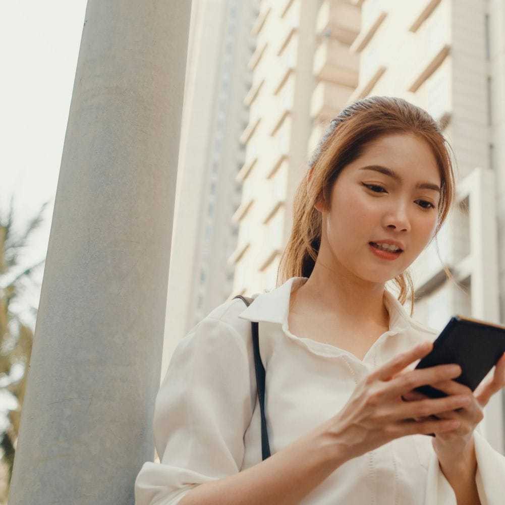 Young woman outdoors, smiling while using her smartphone, with tall buildings and palm trees in the background.