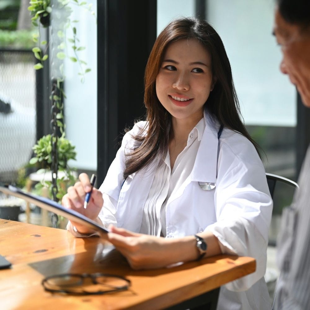 Female doctor smiling while holding a tablet and speaking with a patient during a consultation at a clinic.