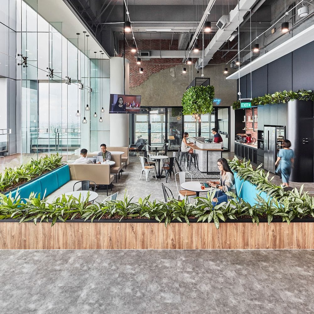 Modern office lounge with people working and having conversations, featuring open seating, greenery, high ceilings, and large windows with natural light.