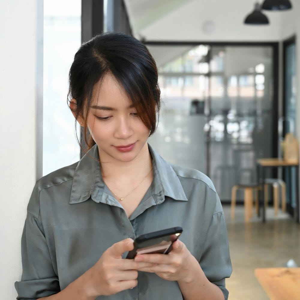 Woman standing indoors and smiling while using a smartphone, in a modern office or café setting.