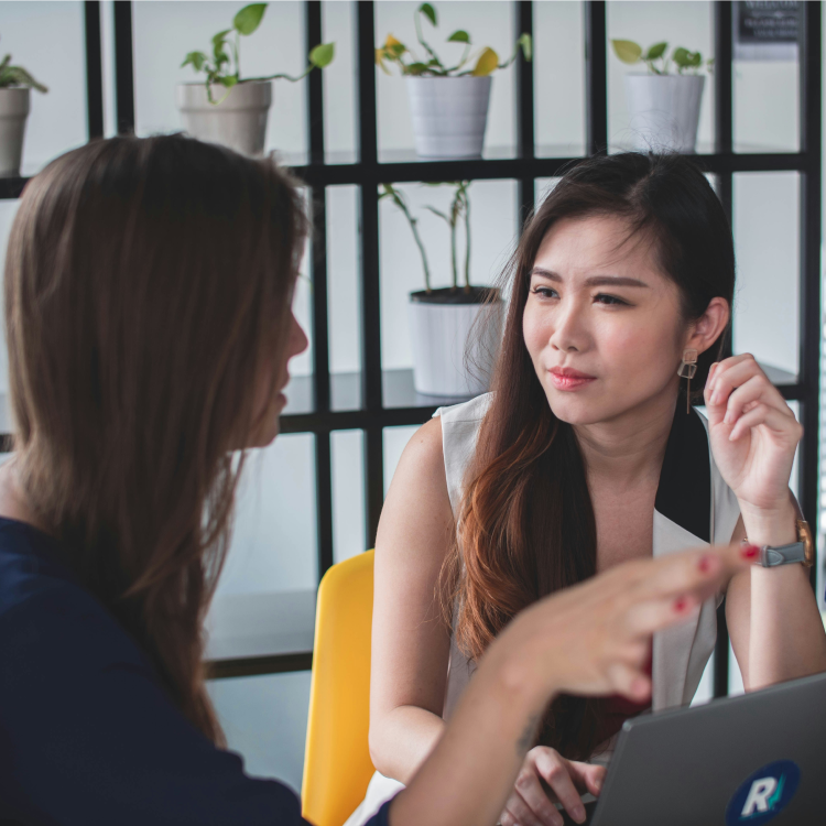 Smiling professional talking on the phone while working on a laptop in a modern office space, with people working in the background.