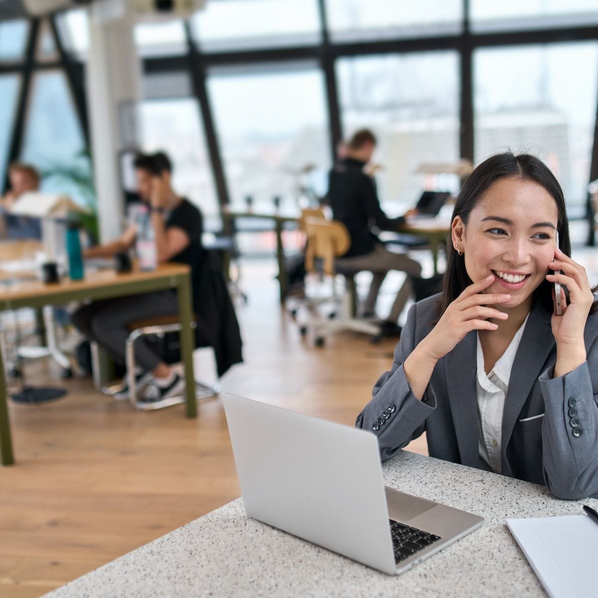 Smiling professional talking on the phone while working on a laptop in a modern office space, with people working in the background.