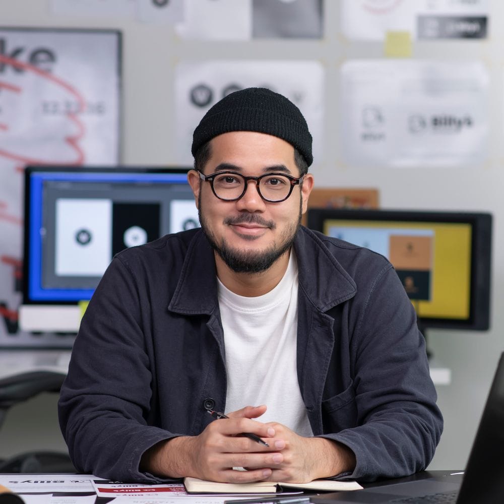 Person sitting at a desk in a creative workspace with design mockups on the wall and computer screens in the background.