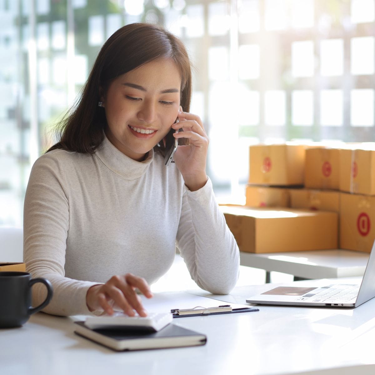 Smiling woman working on a laptop while talking on the phone, sitting at a desk with packaging boxes in the background, suggesting a small business or e-commerce setting.