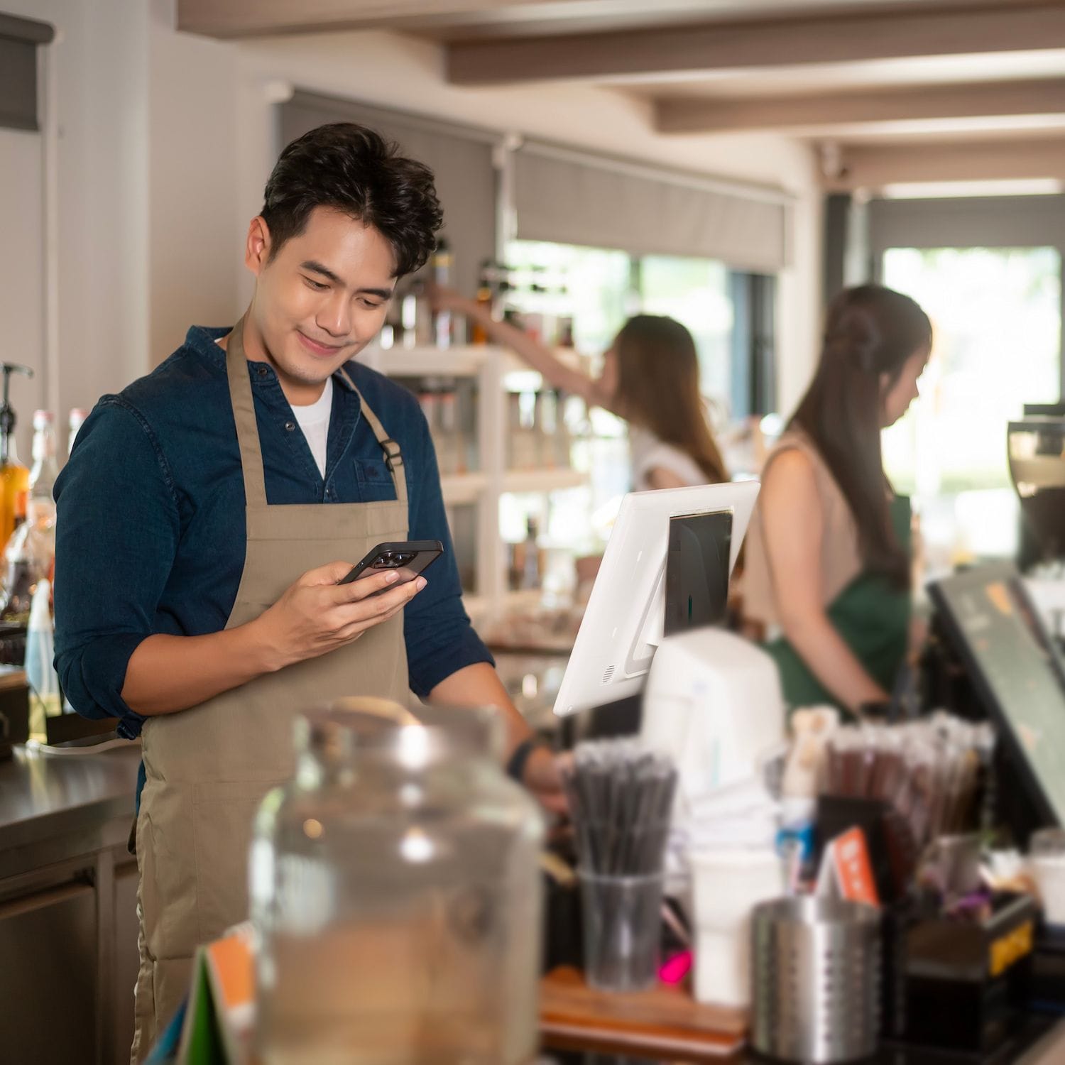 Smiling café worker in an apron using a digital tablet behind the counter, with customers and coffee equipment in the background, representing a small business setting